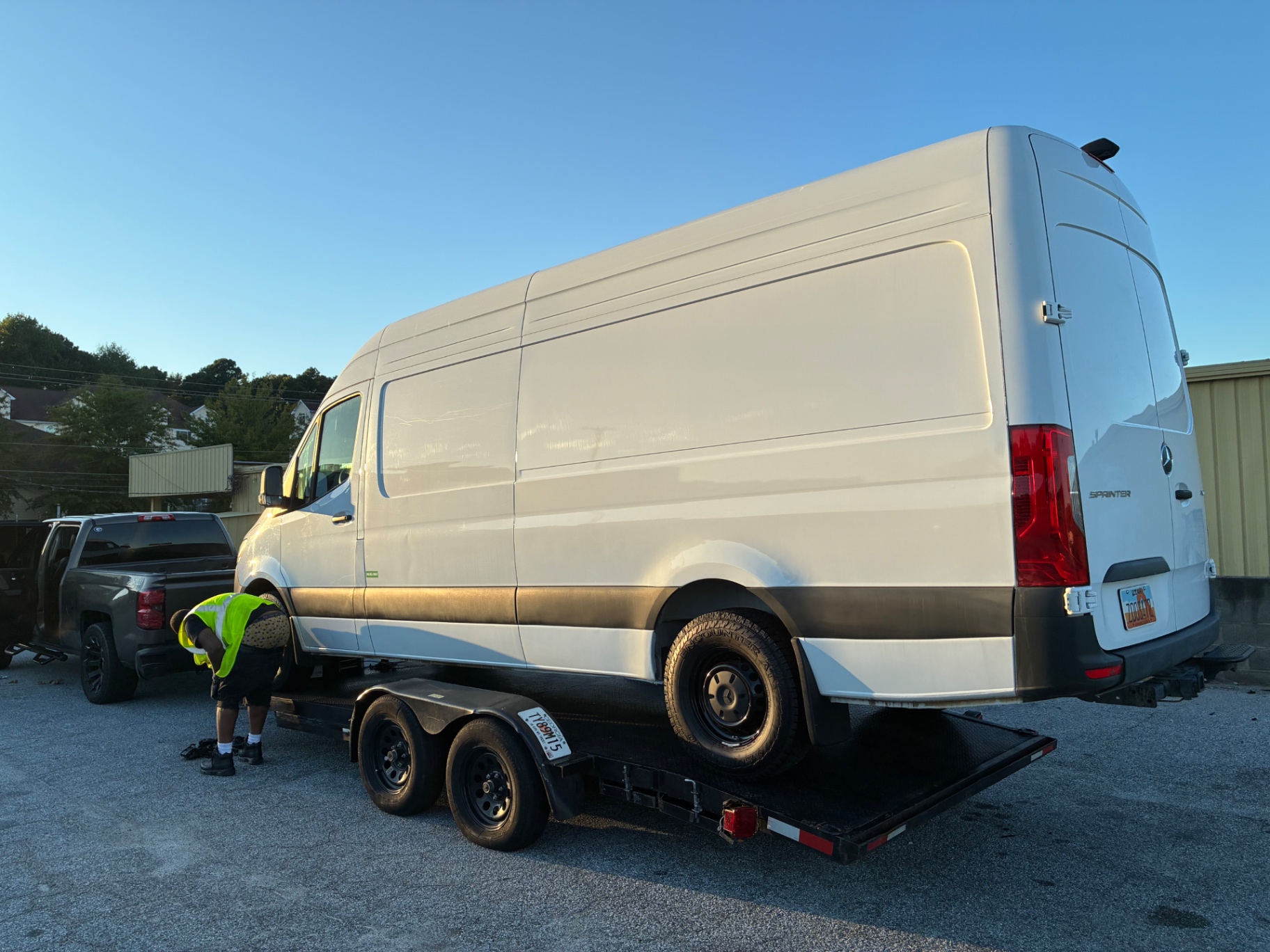Mercedes-Benz Sprinter van loaded on a dual-axle flatbed trailer while a technician secures the tie-down straps