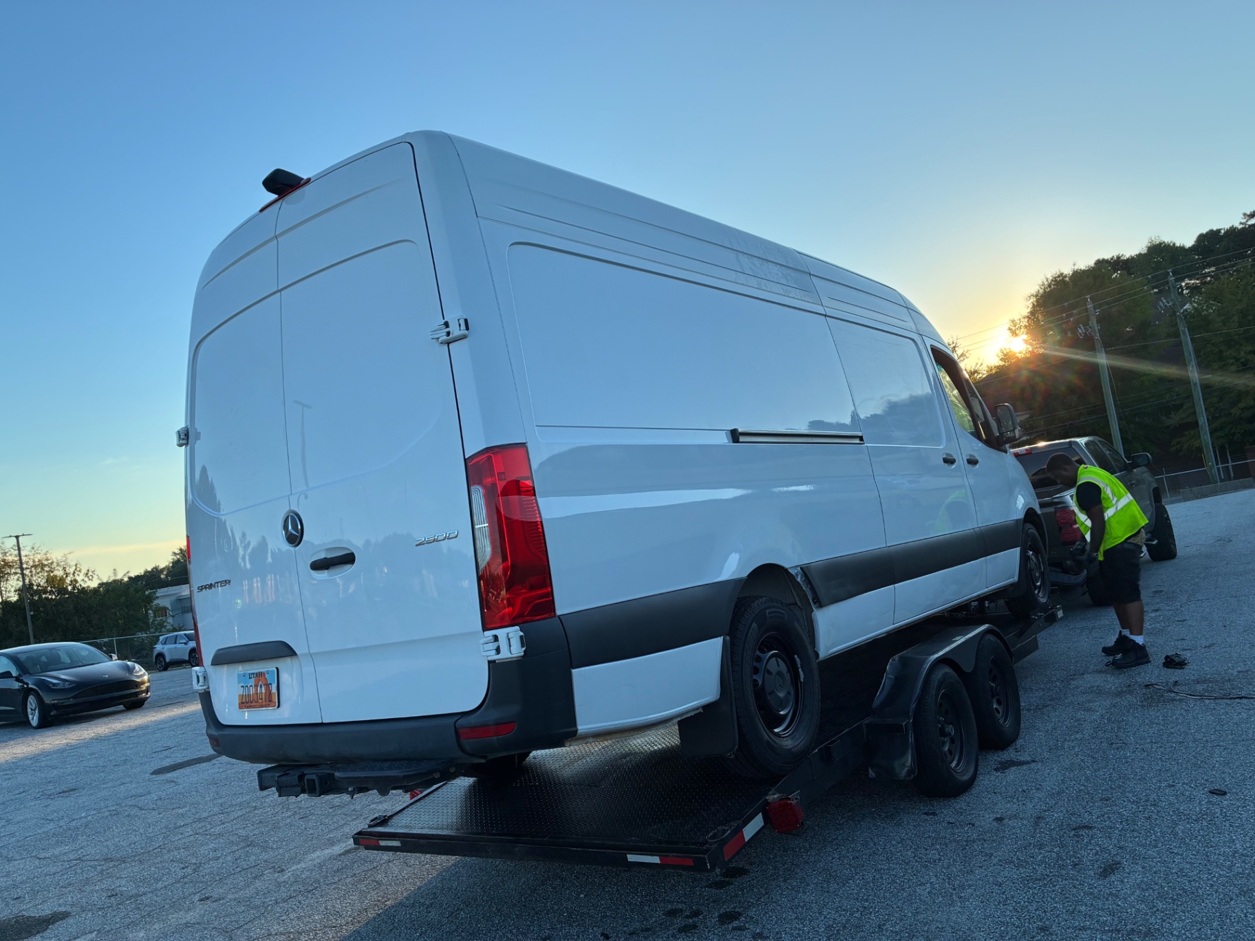 Sprinter van on a flatbed trailer at sunset with a tow technician standing by the pickup truck