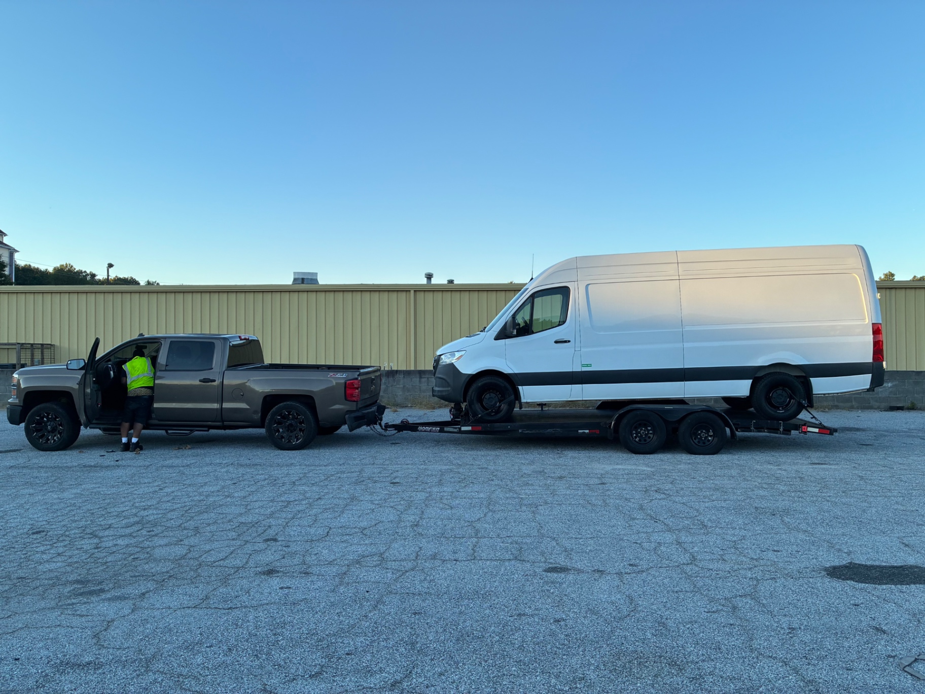 Full side view of a white Sprinter van towed on a flatbed car-hauler trailer behind a pickup truck